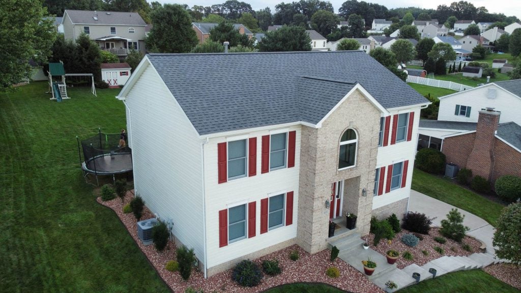 Aerial view of a two-story house with white siding and red shutters. The yard features neatly trimmed grass, landscaped flower beds, a trampoline, and a swing set in the backyard. The neighborhood is surrounded by green trees and similar homes.