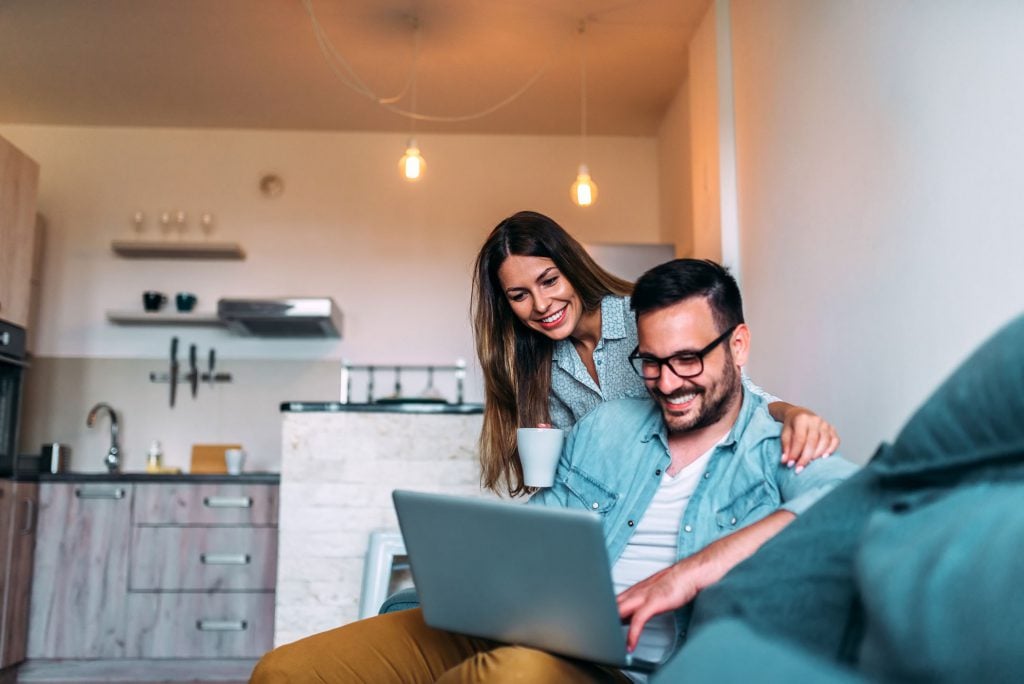 A couple sits on a couch in a modern living room, smiling and looking at a laptop. The woman holds a mug, with her arm around the man, who is wearing glasses and holding the computer on his lap. Warm lighting and a kitchen are in the background.