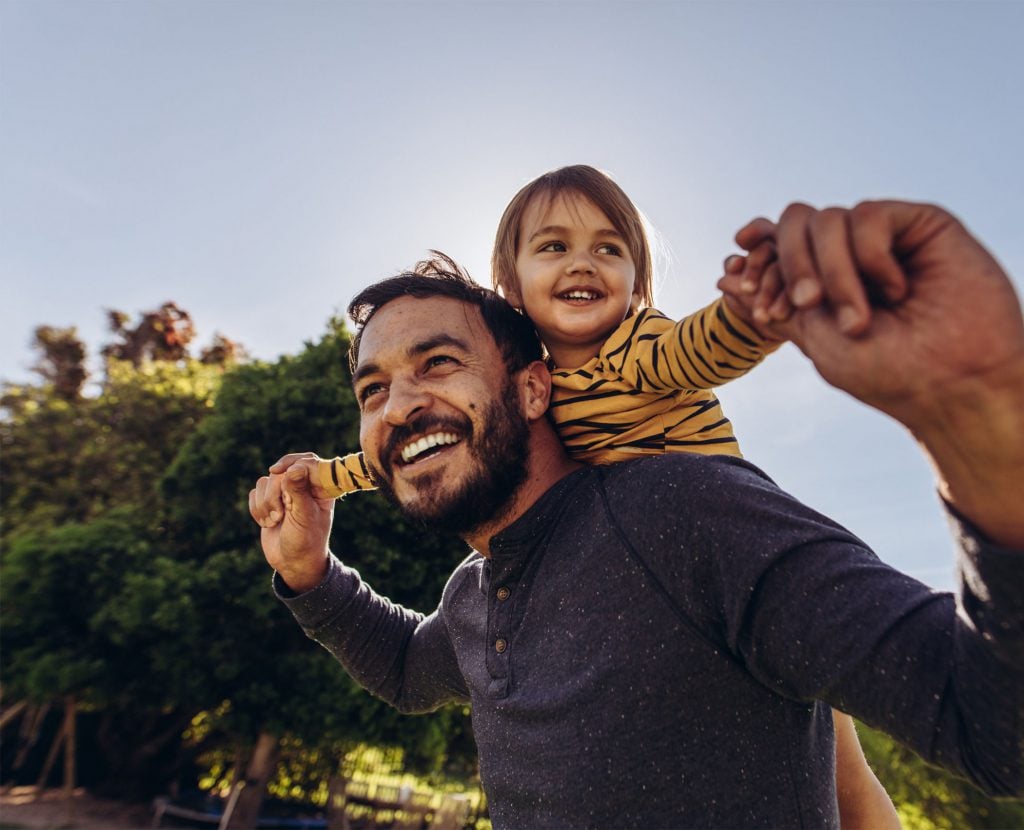 A man with a beard joyfully gives a young child a piggyback ride outdoors. Both are smiling widely. The child is wearing a yellow striped shirt, and the man is in a gray shirt. Green trees and a clear blue sky are in the background.