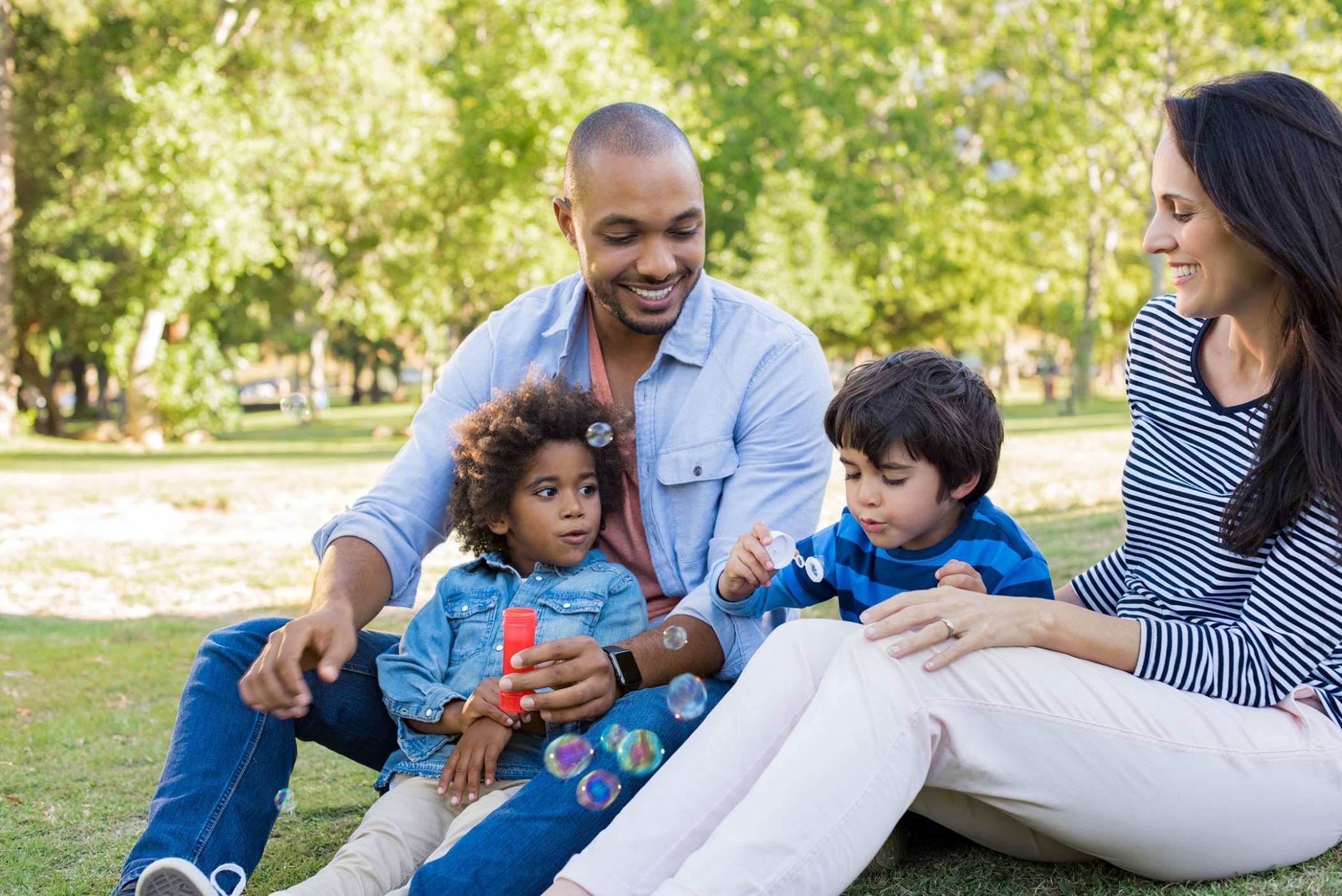 A family of four sits on the grass in a park. Two children, one blowing bubbles, are with two adults. The background features green trees and a sunny atmosphere. Everyone is smiling and dressed in casual attire.