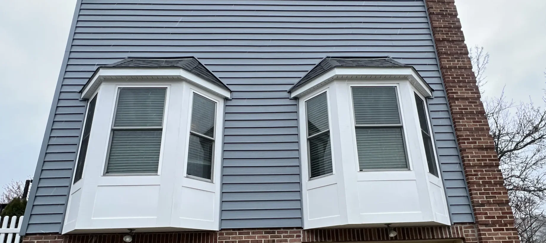 The image shows two white bay windows on the upper floor of a house. The house exterior features gray siding and red brick accents. The windows have blinds and are against a cloudy sky backdrop.