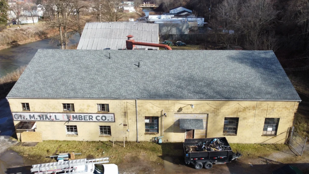 Aerial view of an old, large rectangular building labeled "G.M. Hall Lumber Co." with a gray roof and faded yellow walls. A dumpster filled with debris is in front, surrounded by patches of grass and adjacent buildings. Trees and a creek are visible behind.