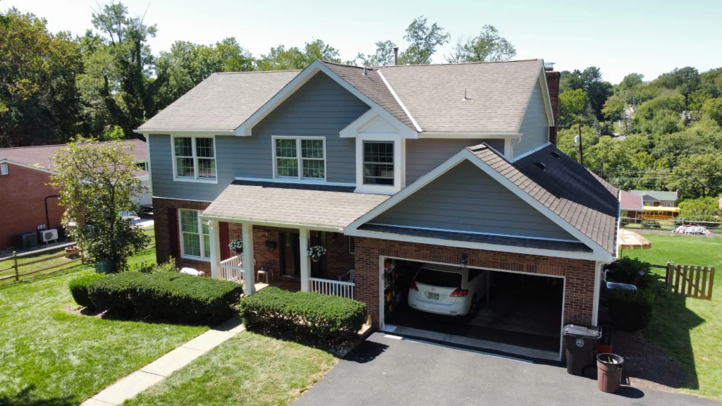 A two-story suburban house with blue and brick exterior, white trim, and a gray roof. It has a neatly maintained lawn, a driveway, and an open garage with a white car inside. Bushes line the walkway to the front porch. Trees are in the background.