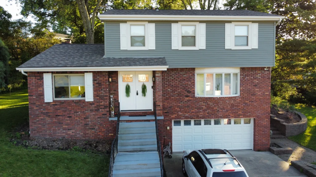 A two-story brick and vinyl siding house with a white garage door and front steps leading to the entrance. There's a white car parked in the driveway and lush greenery surrounds the home.