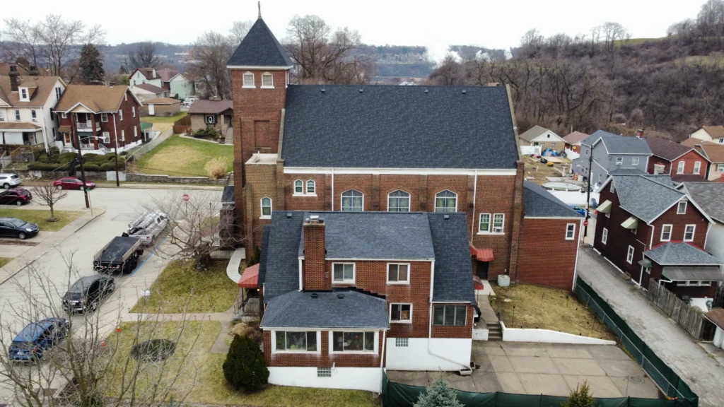 Aerial view of a large brick building with a tower, resembling a church, in a neighborhood. Several smaller homes surround it, and bare trees dot the landscape. The sky is overcast, and the scene conveys a chilly, late-autumn atmosphere.