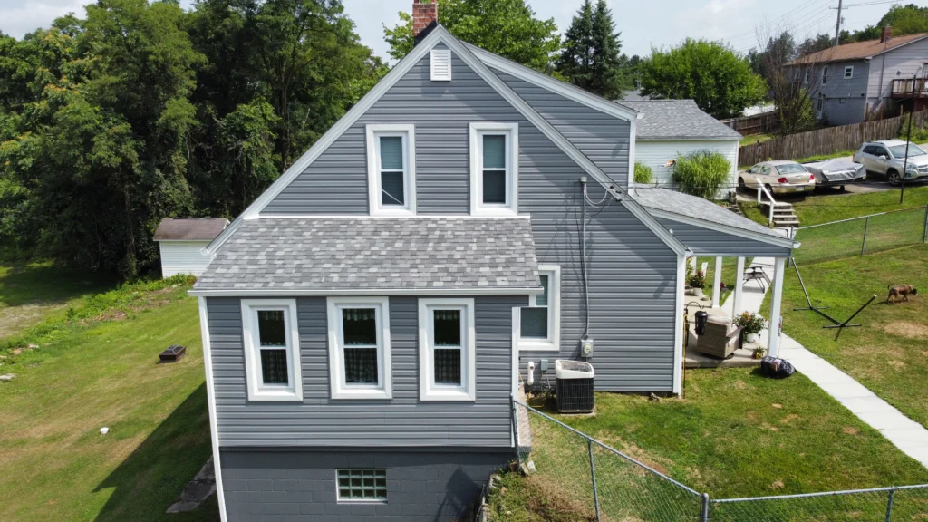 A gray two-story house with a gabled roof, featuring several white-framed windows. The house is surrounded by a green lawn, a chain-link fence, and trees. A small porch with outdoor furniture is visible on the right side.