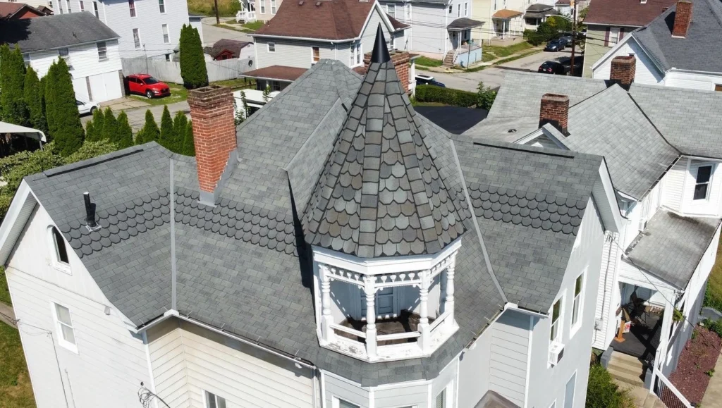 Aerial view of a gray-shingled house with a distinctive turret featuring scalloped shingles and a wooden railing. The house has a chimney and is surrounded by similar residential buildings. A red car is parked nearby.