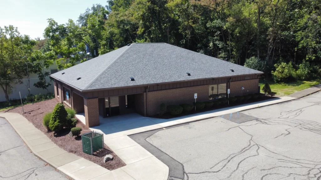 A single-story brick building with a dark gray roof sits amid a paved parking lot with marked spaces. Surrounding it are trees and shrubs. The building has a covered entrance and several windows along its walls.