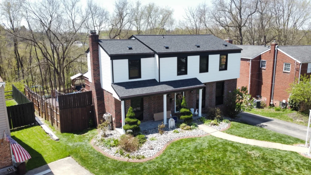 A two-story house with black and white exterior features a neatly landscaped front yard with shrubs and stones. A pathway leads from the driveway to the entrance. Trees and neighboring houses are visible in the background.