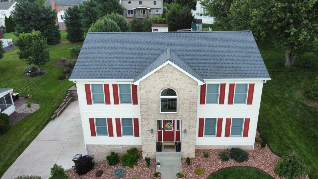Aerial view of a two-story house with beige siding, red shutters, and a brick facade around the entrance. The roof is dark gray, and the yard is landscaped with bushes and lawn. A driveway is on the left side of the house.