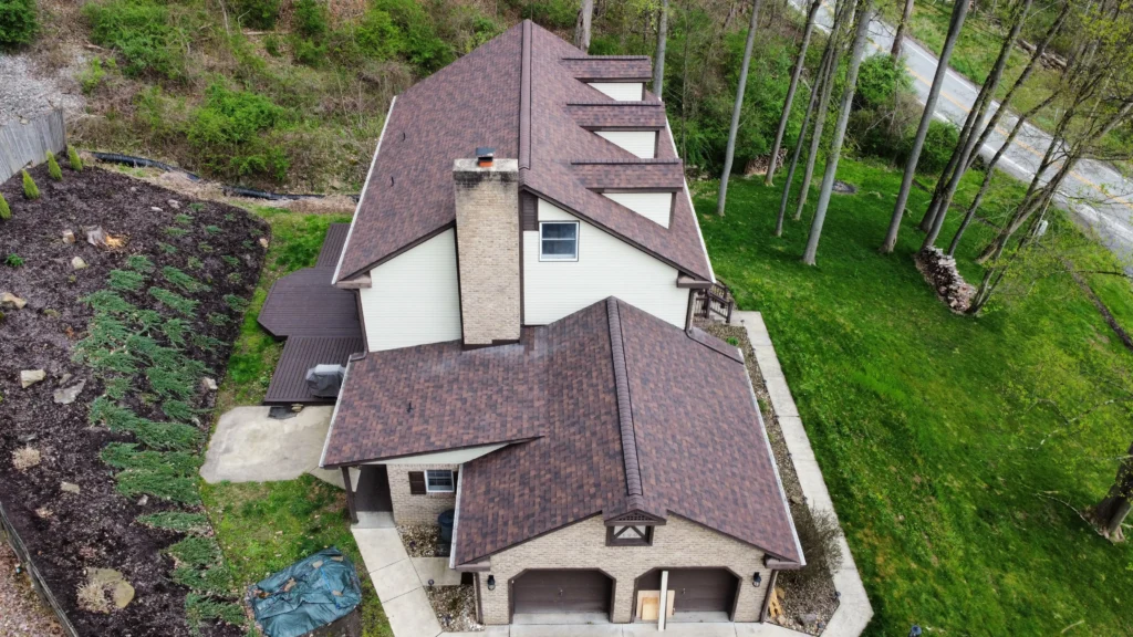 Aerial view of a two-story house with a brown roof surrounded by greenery. The house has multiple skylights and a central chimney. A driveway and landscaped garden with shrubs are adjacent to the structure. Trees line the property boundary.