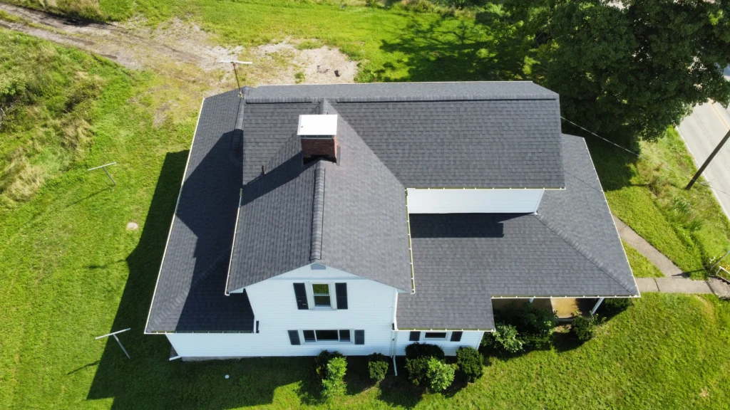 Aerial view of a white two-story house with a dark roof, surrounded by a lush green lawn. The house has a chimney and several windows, with trees and a road visible in the background.