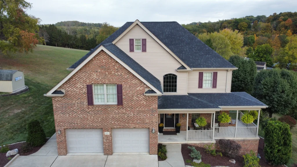 A two-story house with a red brick exterior on the lower level and beige siding on the upper level. It features a two-car garage, a front porch with hanging plants, and is surrounded by trees and a grassy lawn.