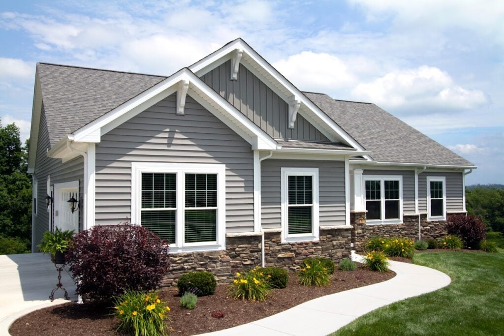 A modern single-story house with gray siding and stone accents. It has multiple windows and a gable roof. The front yard features well-maintained landscaping with bushes and yellow flowers. A curved concrete path leads to the entrance.