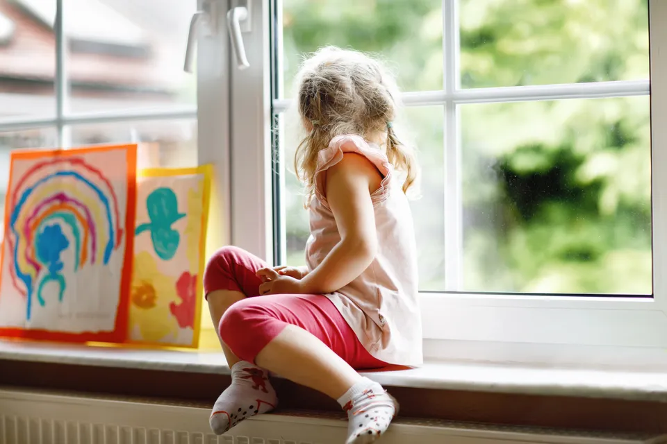 Little girl by window with artwork next to her on rainy day looking out the window.