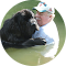 A man in a cap stands in water, holding a large black dog. Both seem calm and content in the sunny outdoor setting.