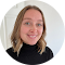 A person with shoulder-length blonde hair smiles at the camera. They are wearing a black turtleneck sweater. The background is a light-colored wall.