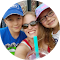 A woman wearing sunglasses smiles while holding a drink with a straw. She is flanked by two children wearing hats and glasses. They are outdoors on a sunny day.