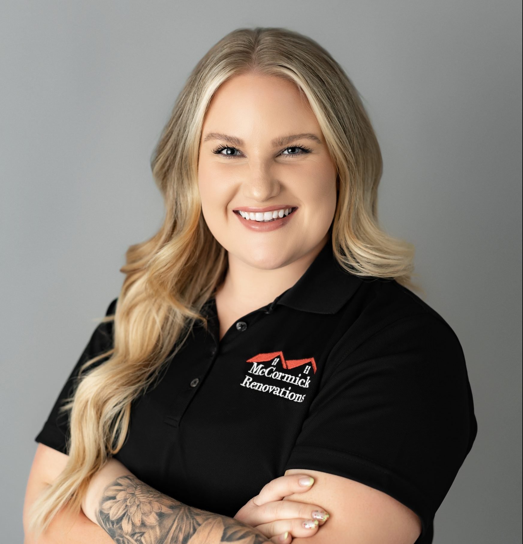 A smiling person with long blonde hair poses with arms crossed, wearing a black polo shirt with a company logo that reads 