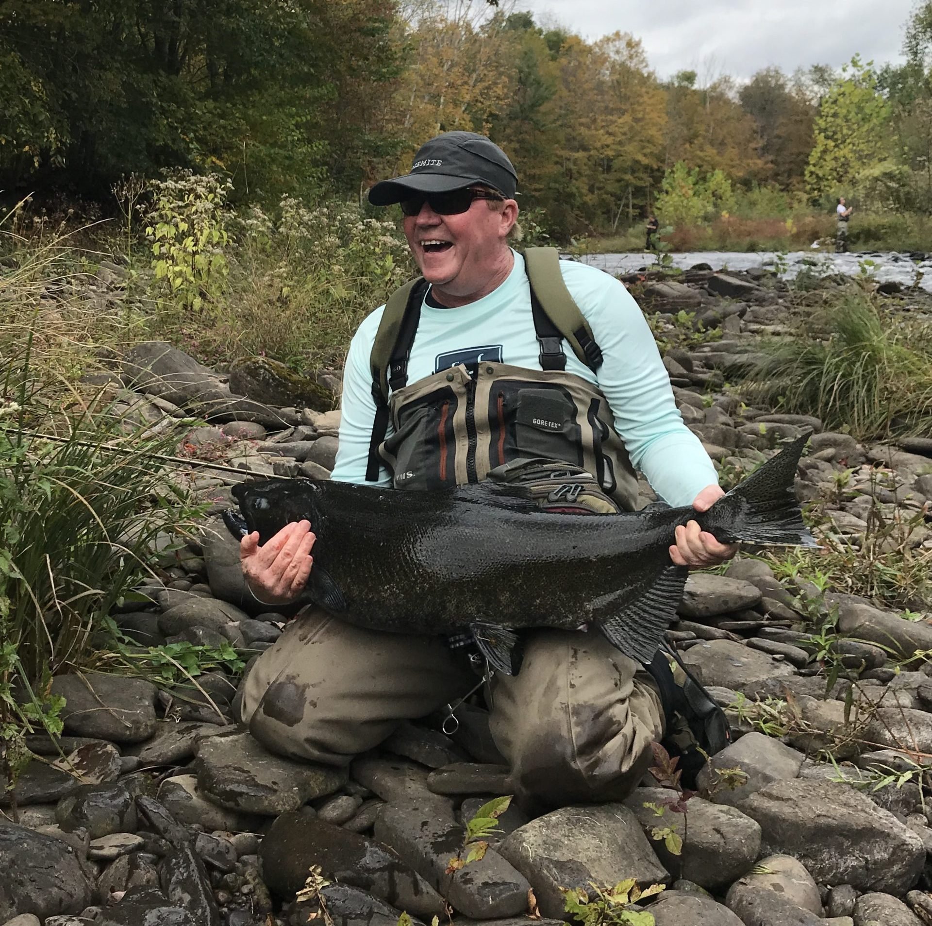 A man wearing sunglasses and waders is sitting on rocks by a river, holding a large fish with a joyful expression. The background features trees with autumn foliage.