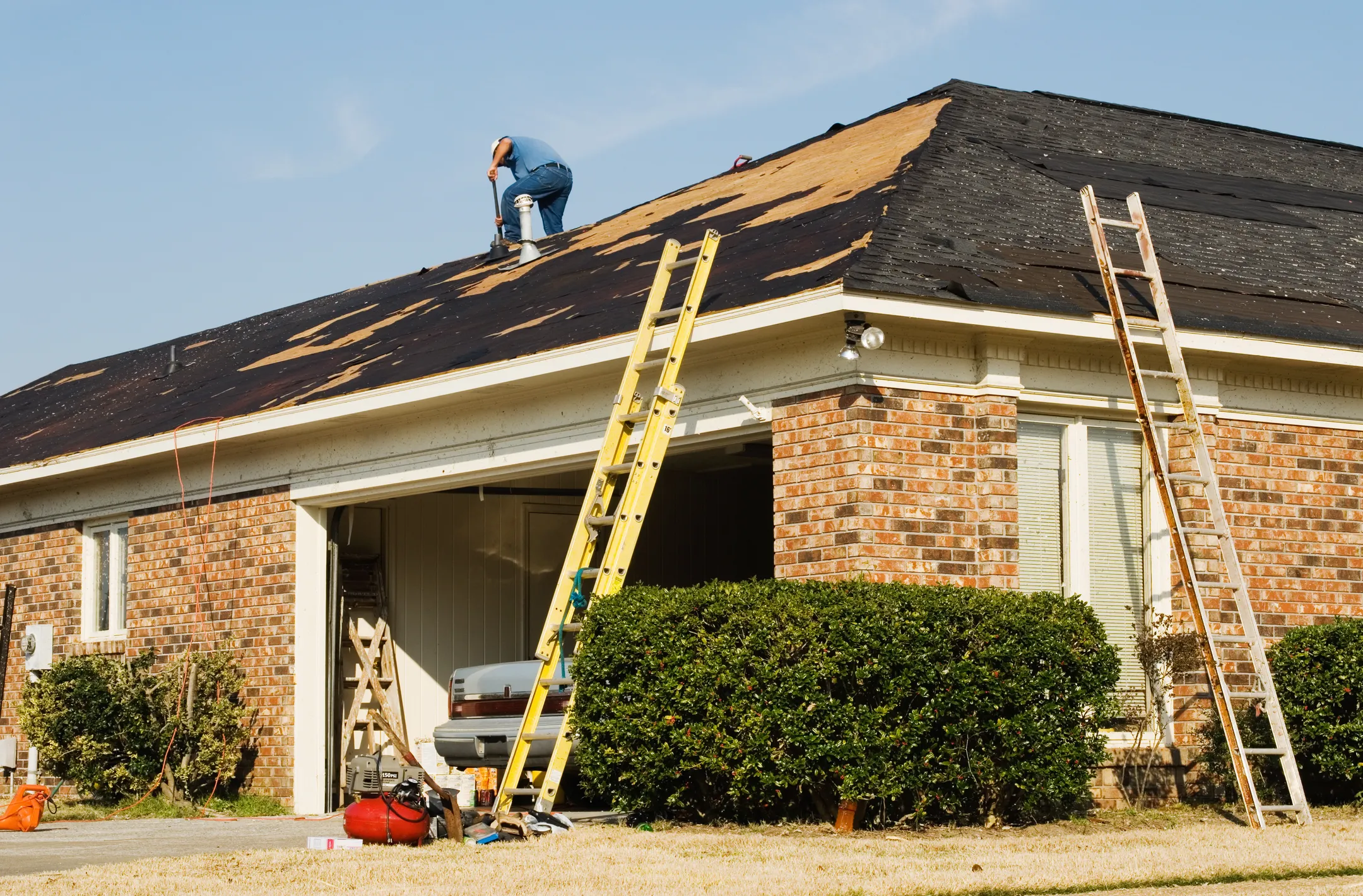 A person tackles a DIY roof repair on a brick house under renovation. Two ladders are propped against the roof, and the garage door is open, revealing tools and equipment inside. A blue sky serves as the backdrop for this hands-on project.