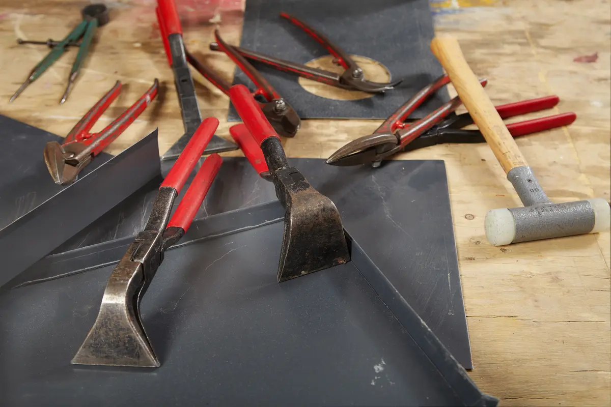 A collection of metalworking tools, including hammers, pliers, and clamps with red handles, are arranged on a wooden workbench. Sheets of metal and measuring tools are also visible, suggesting a workspace for craftsmanship.