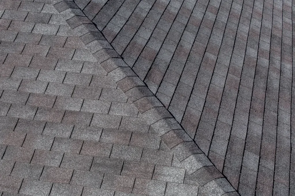 Close-up of a roof with overlapping asphalt shingles. The shingles are gray and aligned in a uniform pattern, with a ridge running diagonally through the image, creating a symmetrical design.