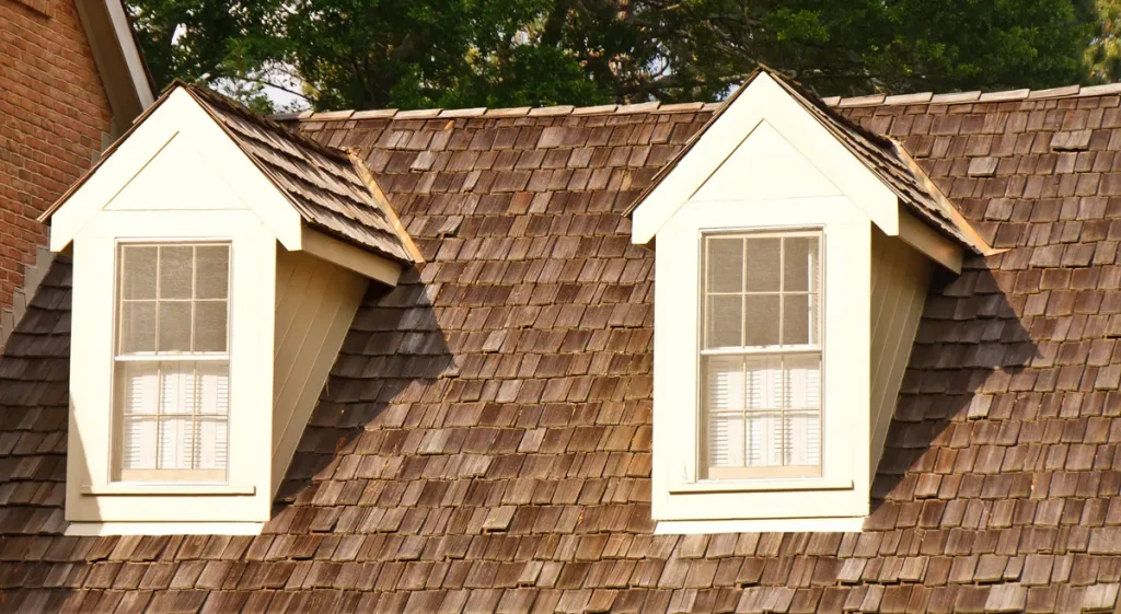 A close-up of a wooden shingle roof with two dormer windows. The windows have cream-colored frames and are topped with gabled roofs. Trees are visible in the background.