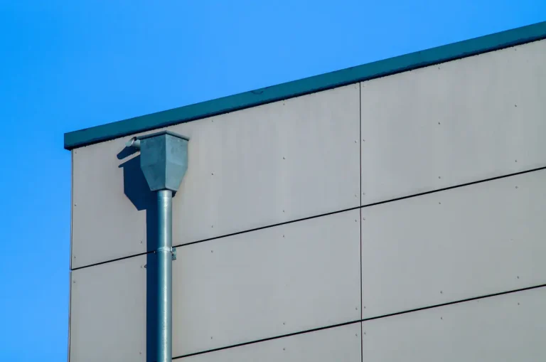 A minimalist image shows the corner of a modern building with a smooth beige facade and a steel drainpipe. The clear blue sky provides a contrasting background, highlighting the building's geometric design.