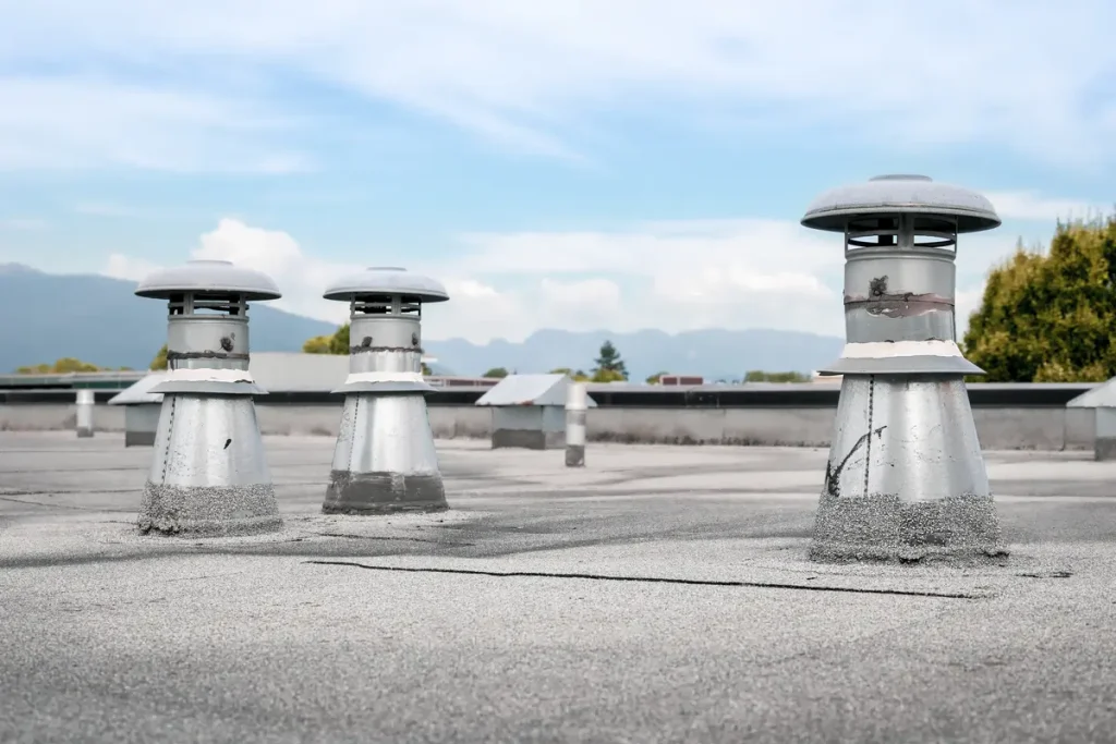 Three metallic chimney vents protrude from a flat rooftop against a backdrop of blue sky and distant mountains. The vents are cylindrical with vents near the top, resembling small towers on the building's surface.