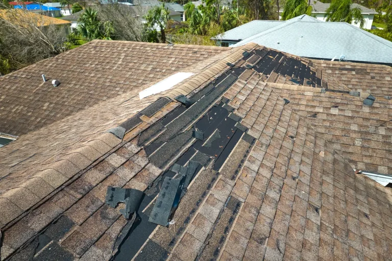 Aerial view of a roof with extensive shingle damage. Several shingles are missing, exposing black underlayment. Surrounding the roof are green trees and other houses with intact roofs in the neighborhood.