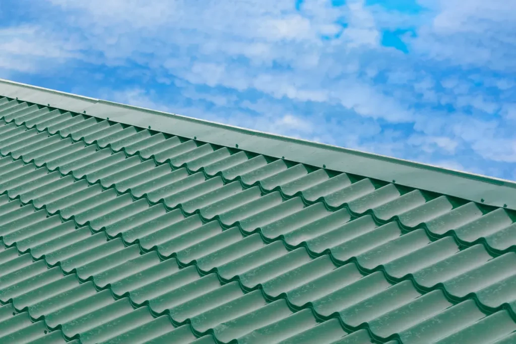 Close-up of a green metal roof with a patterned texture, sloping diagonally across the image. The background features a blue sky with scattered white clouds.