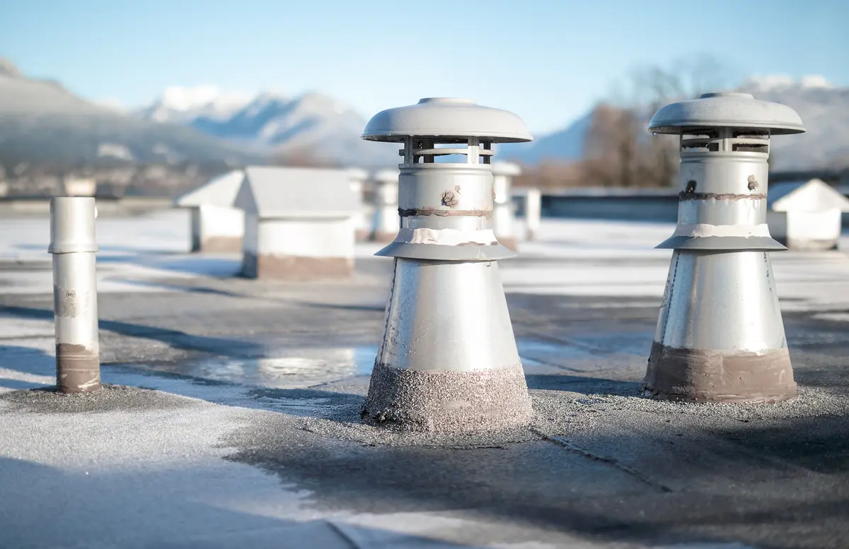 Frost-covered rooftop with several metal chimneys and vents. Snow-capped mountains and blurred trees are in the background under a clear blue sky.