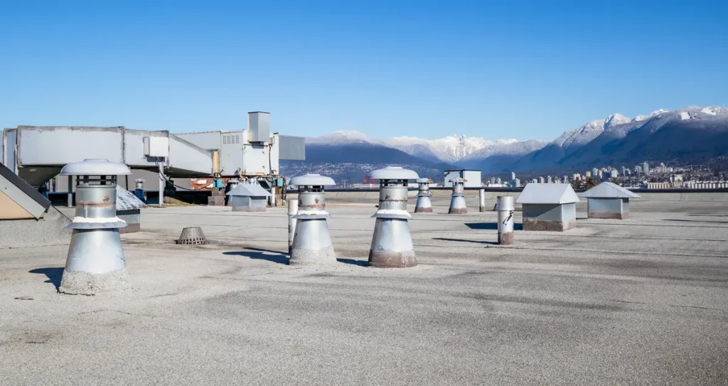 A commercial rooftop with multiple ventilation units and air conditioning systems. In the background, snow-capped mountains are visible under a clear blue sky.