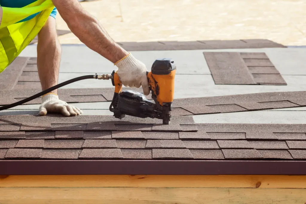 A construction worker wearing gloves uses a nail gun to install asphalt shingles on a roof. The worker's arm and the tool are in focus, with the roof surface clearly visible, showcasing a step in the roofing process.