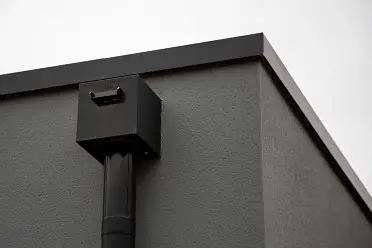 A close-up of a modern building corner with minimalist architectural design. The building features a smooth gray surface with a black drainage pipe running vertically, ending in a rectangular outlet near the top. The sky is overcast.
