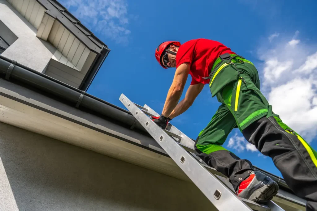A worker in a red shirt and helmet climbs a ladder leaning against a building. The sky is clear and blue, and the worker is wearing safety gear, including green pants and gloves, while inspecting or working on the roof area.