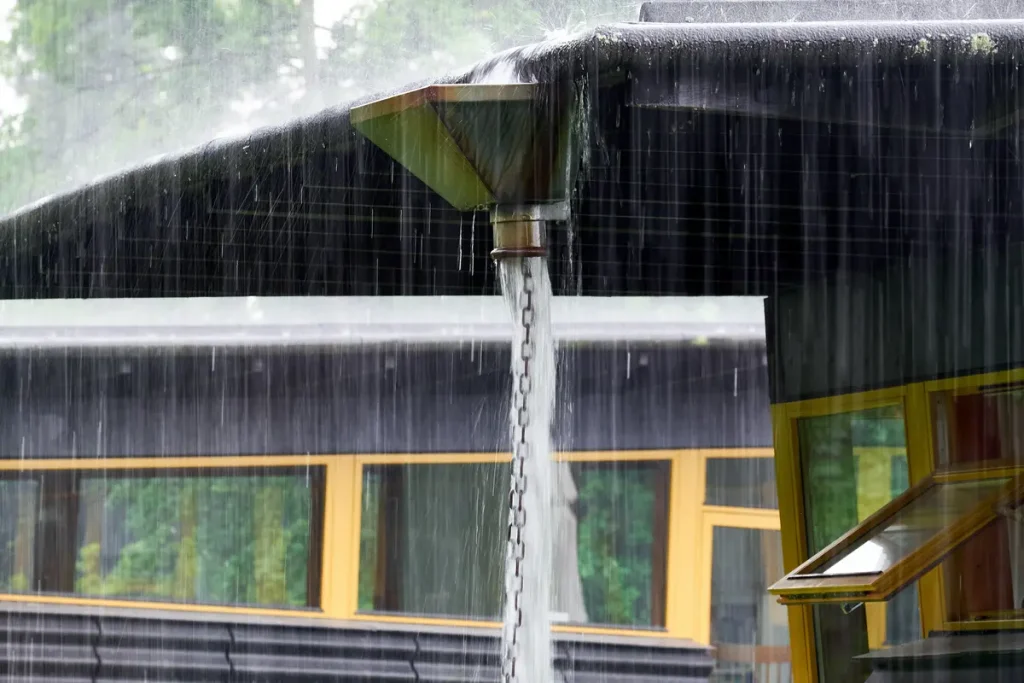 A chain downspout guides rainwater from a slanted roof, creating a curtain of water. A building with large yellow-framed windows and blurred green trees are visible in the background.