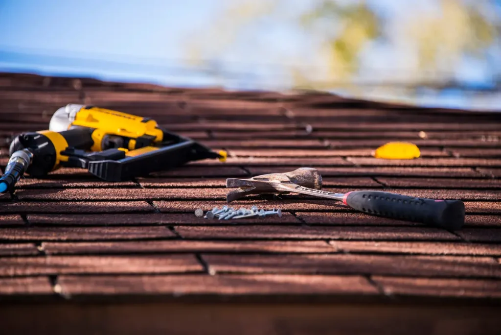 A rooftop with a hammer, nails, and a nail gun lying on the brown shingles. The tools suggest roofing work or repairs under a clear sky.