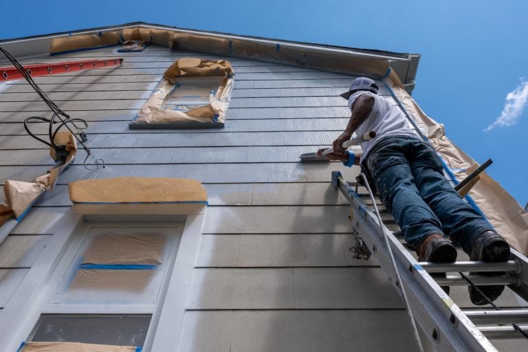 Person on ladder wearing a hat and jeans, painting the exterior of a house. The house is covered with protective paper around windows and edges. Clear blue sky in the background.