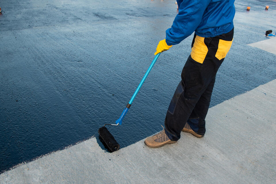 Person applying a black waterproofing coating to a concrete surface with a roller. The individual is wearing a blue jacket, yellow gloves, and dark pants. The scene captures a partial view of the person's lower body.