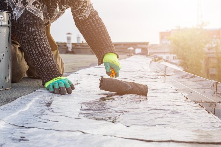 A person wearing bright green gloves and a patterned sweater applies waterproofing sealant with a roller brush to a flat roof. The sun shines in the background, casting light over the urban setting.