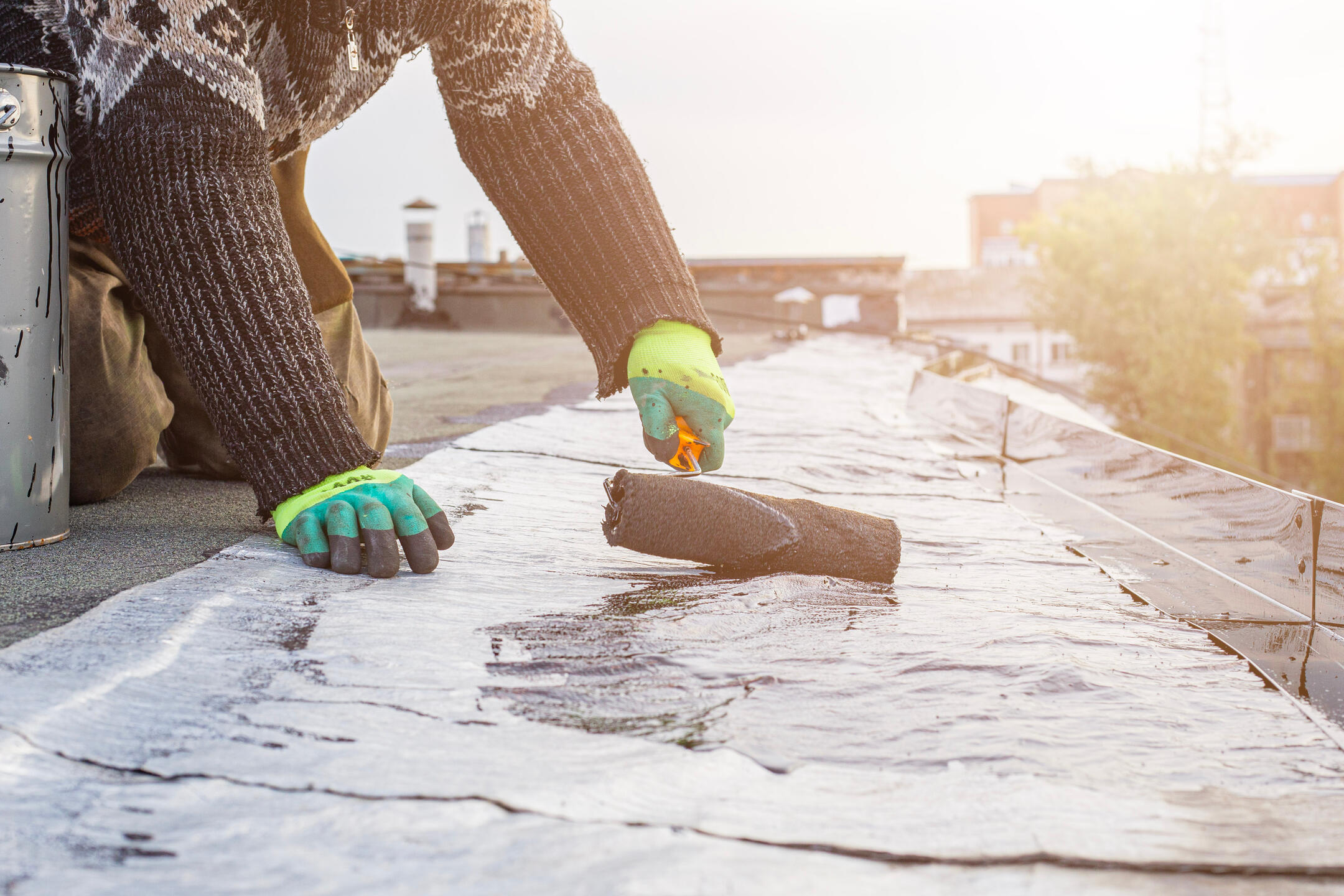A person wearing bright green gloves and a patterned sweater applies waterproofing sealant with a roller brush to a flat roof. The sun shines in the background, casting light over the urban setting.