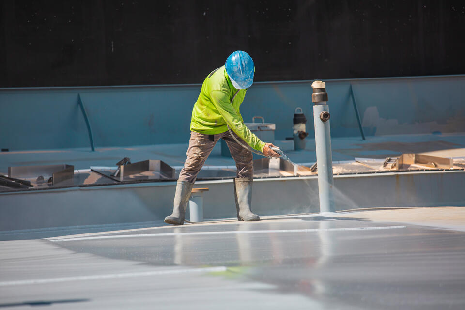 A construction worker in a bright green jacket and blue hard hat sprays sealant on a large flat surface, possibly a rooftop or floor. The area is surrounded by various construction materials and equipment.