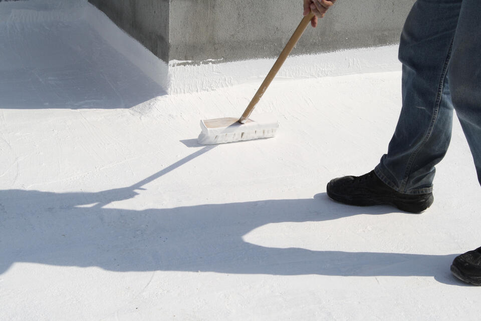 A person uses a long-handled brush to apply white coating to a flat roof. The ground and surrounding walls are also coated in white. The individual's shadow is visible on the surface. They wear jeans and dark shoes.