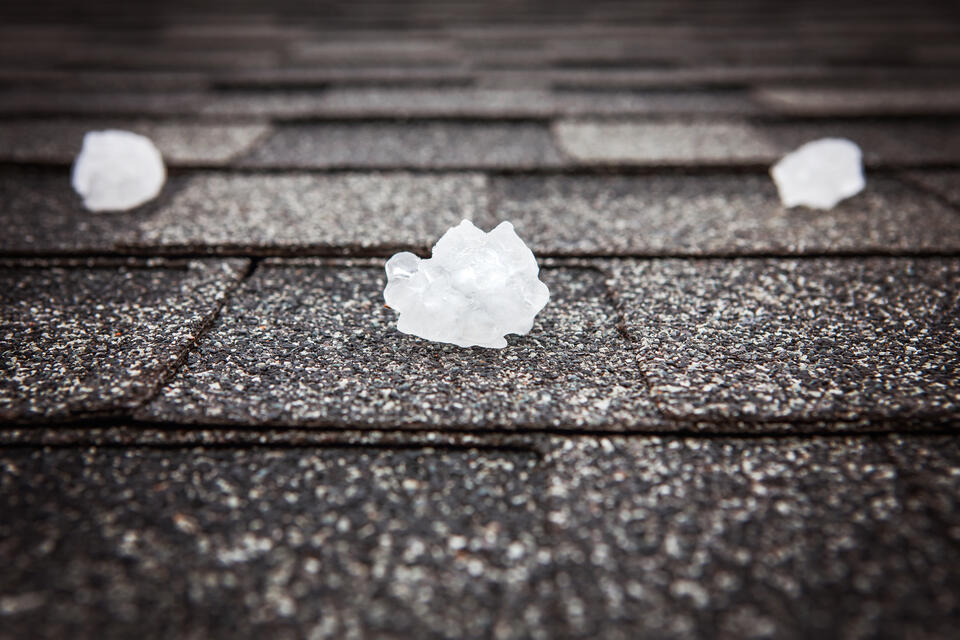 Close-up of large hailstones resting on a dark gray shingled roof. The surface of the roof is textured, and the hailstones appear irregular and translucent, indicating recent or ongoing hailstorm activity.