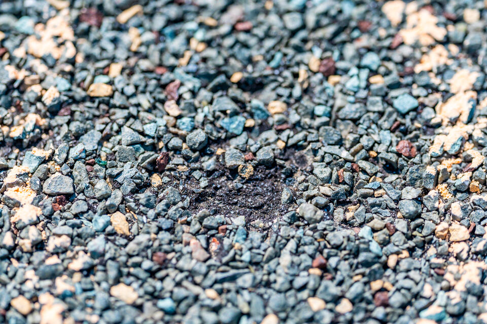 Close-up of a textured surface of a blue-gray and tan asphalt shingle, showing a slight indentation in the center. The granular particles vary in color, creating a speckled appearance.