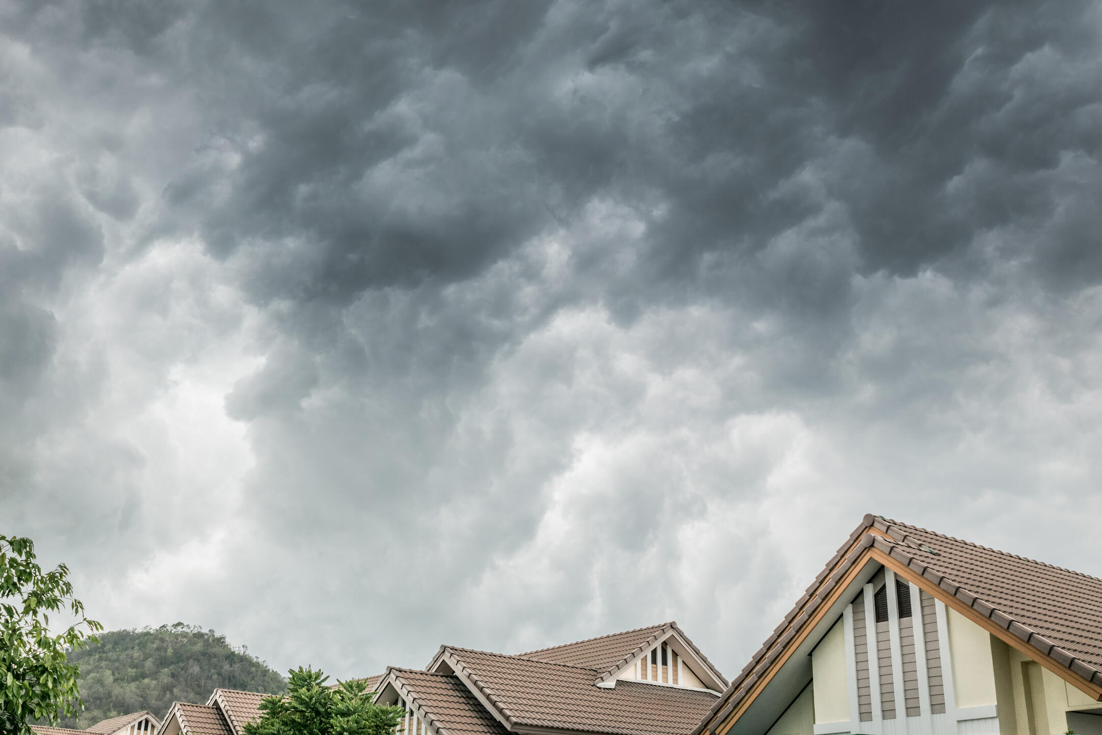 Stormy clouds loom over a row of houses with brown tiled roofs. The sky is dark and ominous, suggesting an approaching storm. A few trees are visible, and hills can be seen in the distance.