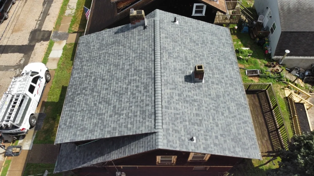Aerial view of a house in Monroeville with a new gray shingle roof, showcasing multiple chimneys. The surrounding area includes a white vehicle on the street, neighboring buildings, and a small garden with a wooden deck.