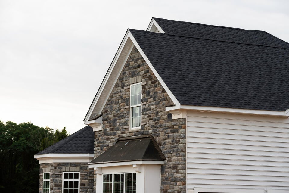 A partial view of a house with a mix of stone and white siding, featuring a dark shingle roof. Multiple windows are visible, and the structure is set against a backdrop of a clear sky and some trees.